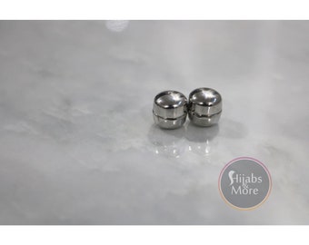 May include: Two silver magnetic hijab pins on a white marble surface.