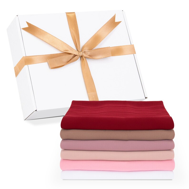 May include: A white gift box with a gold ribbon tied in a bow. The box is sitting on top of a stack of five fabric squares in shades of red, brown, pink, and white.