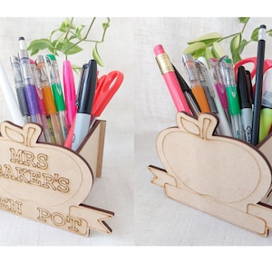 May include: Wooden pen holder shaped like an apple with the text "Mrs. Baker's Pen Pot" on the front. The holder is filled with pens, pencils, and markers.