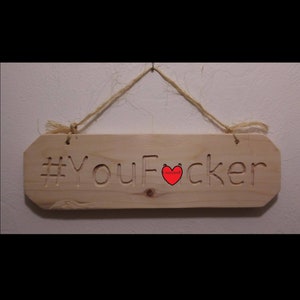Puede incluir: Un letrero de madera con el texto "#YouF*cker" tallado en él. Un corazón rojo está tallado en el letrero entre las palabras "You" y "F*cker". El letrero está colgado de una cuerda.