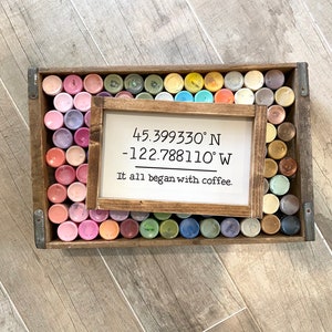 May include: A wooden sign with the coordinates 45.399330&deg; N -122.788110&deg; W and the text "It all began with coffee." The sign is surrounded by a variety of small paint bottles in a wooden crate.