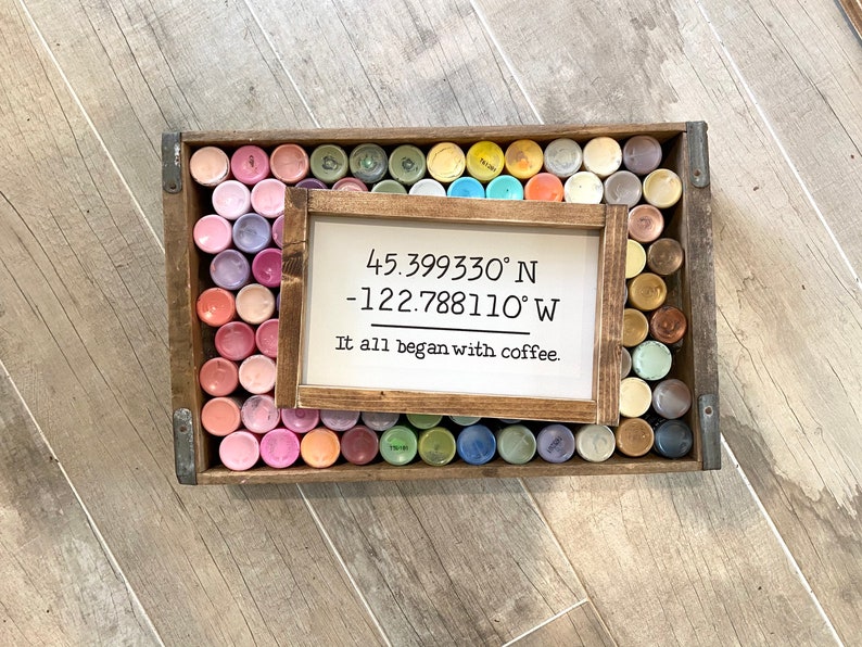 May include: A wooden sign with the coordinates 45.399330&deg; N -122.788110&deg; W and the text "It all began with coffee." The sign is surrounded by a variety of small paint bottles in a wooden crate.