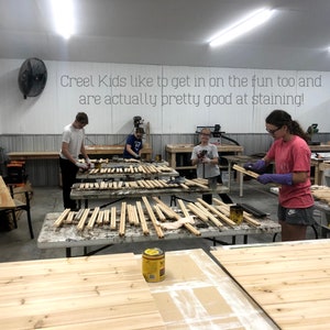 May include: A group of people are working on a woodworking project. They are staining wooden slats with a dark brown stain. The text "Creel Kids like to get in on the fun too and are actually pretty good at staining!" is written on the wall behind them.