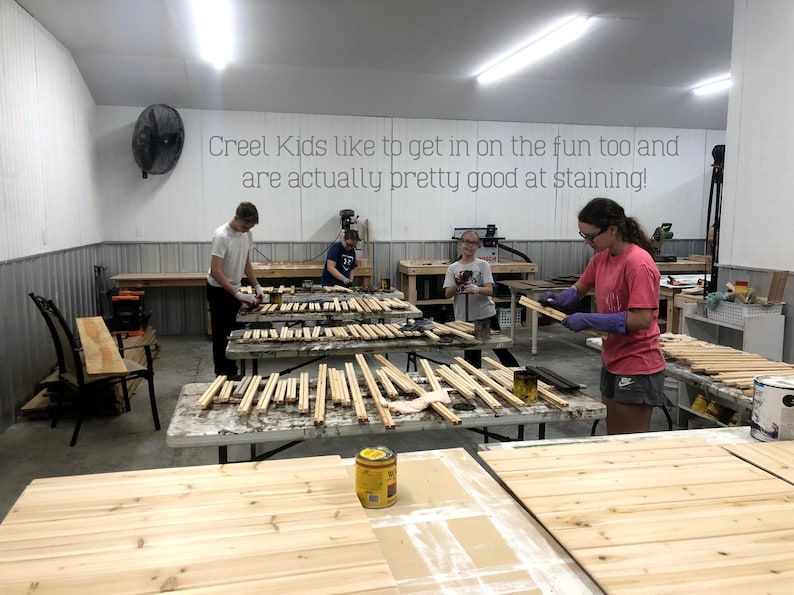 May include: A group of people are working on a woodworking project. They are staining wooden slats with a dark brown stain. The text "Creel Kids like to get in on the fun too and are actually pretty good at staining!" is written on the wall behind them.