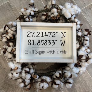 May include: A rectangular white wooden sign with black text reading 27.21472&deg; N 81.85833&deg; W and It all began with a ride. The sign is centered on a circular wreath made of white cotton bolls with brown stems and husks, creating a decorative display.