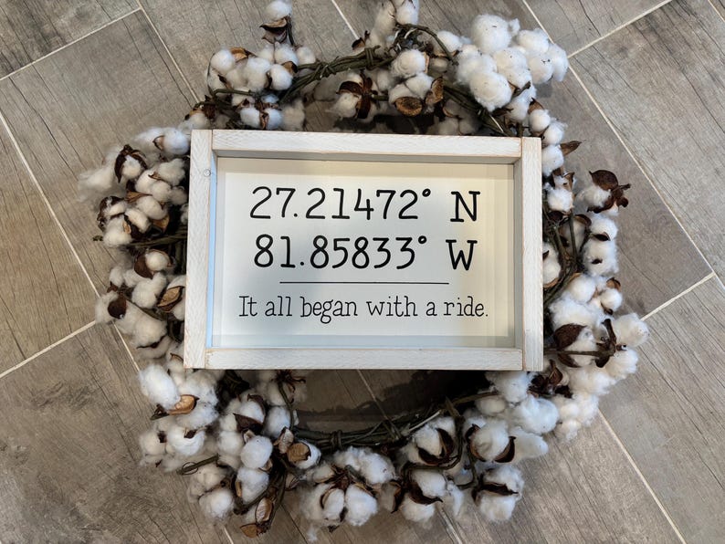 May include: A rectangular white wooden sign with black text reading 27.21472&deg; N 81.85833&deg; W and It all began with a ride. The sign is centered on a circular wreath made of white cotton bolls with brown stems and husks, creating a decorative display.