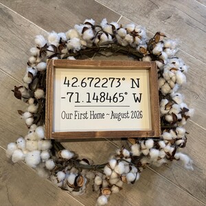 May include: A rectangular wooden sign with a white background and dark brown frame, featuring the text 42.672273&deg; N -71.148465&deg; W and Our First Home ~ August 2026, centered inside a circular wreath made of white cotton bolls and brown twigs.