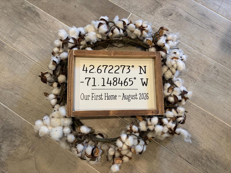 May include: A rectangular wooden sign with a white background and dark brown frame, featuring the text 42.672273&deg; N -71.148465&deg; W and Our First Home ~ August 2026, centered inside a circular wreath made of white cotton bolls and brown twigs.