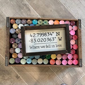 May include: A rectangular wooden sign with a dark brown frame and white background featuring the black text 42.799834&deg; N -83.020363&deg; W and Where we fell in love. The sign rests on a wooden crate filled with many small, colorful paint bottles.