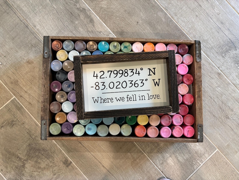 May include: A rectangular wooden sign with a dark brown frame and white background featuring the black text 42.799834&deg; N -83.020363&deg; W and Where we fell in love. The sign rests on a wooden crate filled with many small, colorful paint bottles.