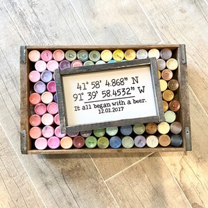 May include: A wooden sign with black text that reads "41&deg; 58' 4.868" N 91&deg; 39' 58.4532" W It all began with a beer. 12.01.2017". The sign is sitting on top of a wooden crate filled with small paint containers.