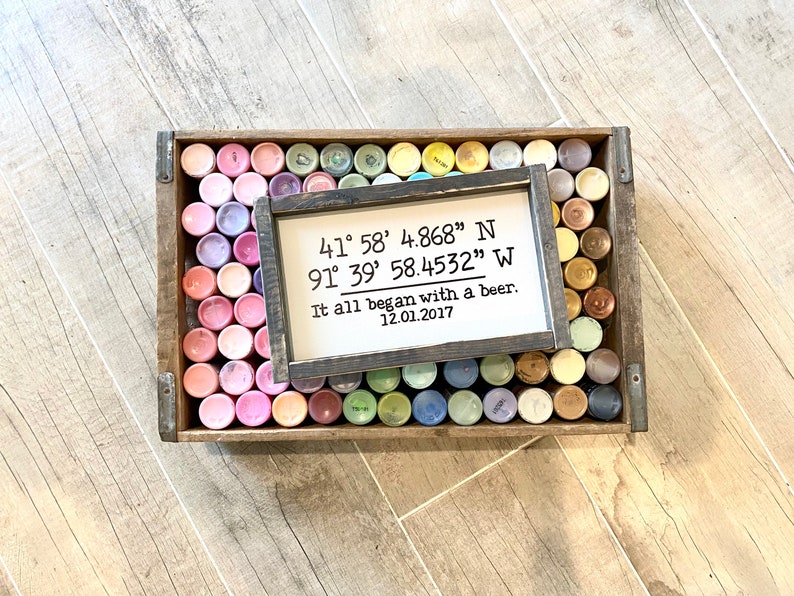 May include: A wooden sign with black text that reads "41&deg; 58' 4.868" N 91&deg; 39' 58.4532" W It all began with a beer. 12.01.2017". The sign is sitting on top of a wooden crate filled with small paint containers.
