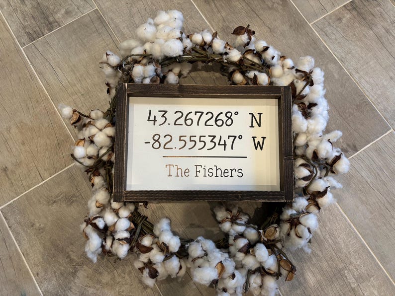 May include: A rectangular wooden sign with a dark brown frame and a white background, featuring the black text 43.267268&deg; N -82.555347&deg; W and The Fishers, centered within a circular wreath made of white cotton bolls and brown woody stems.