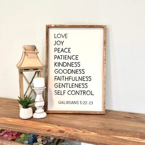 May include: A white wooden sign with black text listing the fruits of the spirit: love, joy, peace, patience, kindness, goodness, faithfulness, gentleness, and self-control. The sign also includes the scripture reference Galatians 5:22-23.