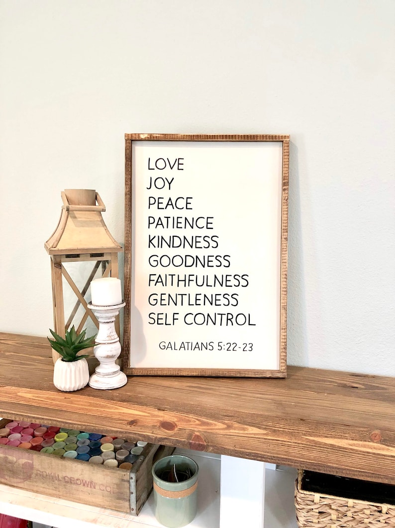 May include: A white wooden sign with black text listing the fruits of the spirit: love, joy, peace, patience, kindness, goodness, faithfulness, gentleness, and self-control. The sign also includes the scripture reference Galatians 5:22-23.