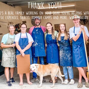 May include: A family of seven wearing blue aprons and working in a woodshop. The sign above them reads "THANK YOU!! THERE'S A REAL FAMILY WORKING ON YOUR SIGN! YOU'RE HELPING WITH OUR COLLEGE EXPENSES, BRACES, SUMMER CAMPS AND MORE. WE APPRECIATE YOU!"