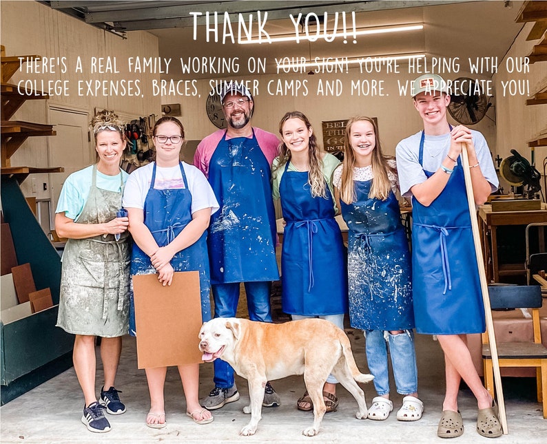 May include: A family of seven wearing blue aprons and working in a woodshop. The sign above them reads "THANK YOU!! THERE'S A REAL FAMILY WORKING ON YOUR SIGN! YOU'RE HELPING WITH OUR COLLEGE EXPENSES, BRACES, SUMMER CAMPS AND MORE. WE APPRECIATE YOU!"