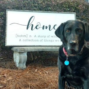 May include: A black Labrador Retriever dog sits in front of a white sign with the word "home" written in cursive. The sign also includes the definition of the word "home" as a story of who and a collection of things we love.