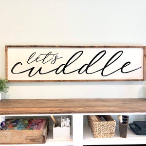 May include: A wooden sign with the text "Let's cuddle" in black cursive script. The sign is framed in a light brown wood frame and is hanging on a light green wall.