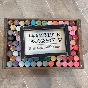 May include: A rectangular wooden crate filled with numerous small, upright paint bottles in various colors. Resting on top of the bottles is a black-framed sign with white text reading 44.447319&deg; N -88.068603&deg; W and It all began with coffee...