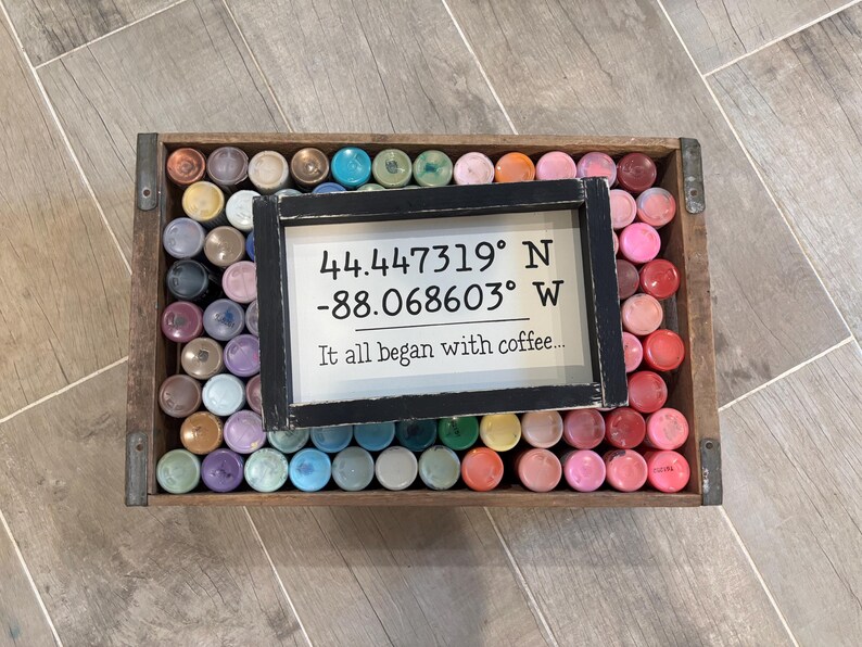 May include: A rectangular wooden crate filled with numerous small, upright paint bottles in various colors. Resting on top of the bottles is a black-framed sign with white text reading 44.447319&deg; N -88.068603&deg; W and It all began with coffee...