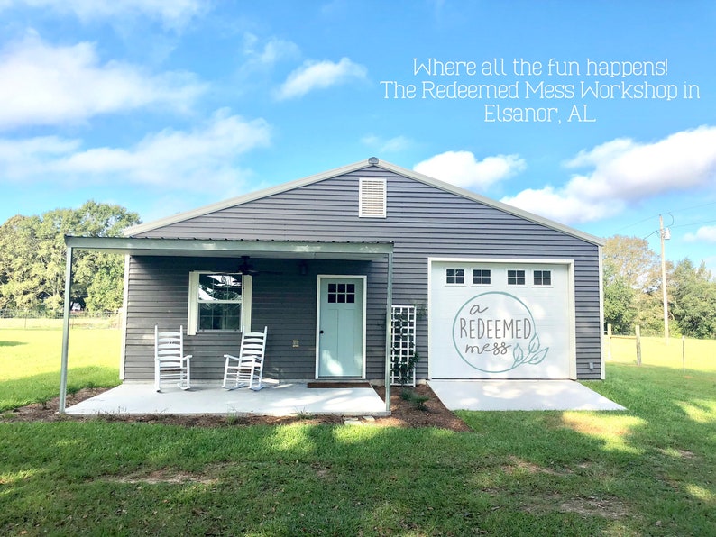 May include: A gray building with a white door and a garage door that says "a redeemed mess" in white lettering. The building has a covered porch with two white rocking chairs.