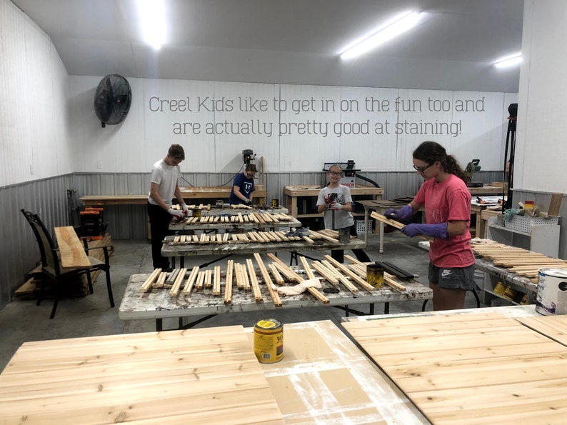 May include: A group of people are working on a woodworking project. They are staining wooden slats with a dark brown stain. The text "Creel Kids like to get in on the fun too and are actually pretty good at staining!" is written on the wall behind them.