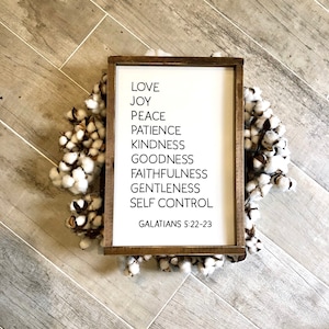 May include: A white wooden sign with black text listing the fruits of the spirit: love, joy, peace, patience, kindness, goodness, faithfulness, gentleness, and self-control. The sign also includes the scripture reference Galatians 5:22-23.