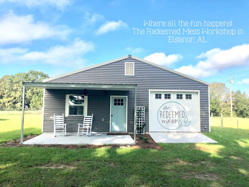 May include: A gray building with a white door and a garage door that says "a redeemed mess" in white lettering. The building has a covered porch with two white rocking chairs.