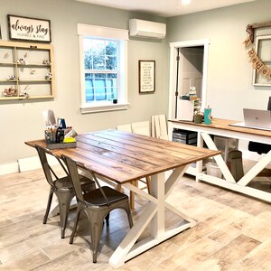 May include: A light green room with two wooden tables, one with white legs and the other with white X-shaped legs. The table with white legs has a laptop computer on it. The table with white X-shaped legs has two metal chairs. The room has a light wood floor and a white ceiling. There is a window on the left side of the room and a door on the right side of the room. There is a sign on the wall that says "Always Stay Humble & Kind".