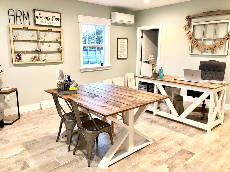 May include: A light green room with two wooden tables, one with white legs and the other with white X-shaped legs. The table with white legs has a laptop computer on it. The table with white X-shaped legs has two metal chairs. The room has a light wood floor and a white ceiling. There is a window on the left side of the room and a door on the right side of the room. There is a sign on the wall that says "Always Stay Humble & Kind".