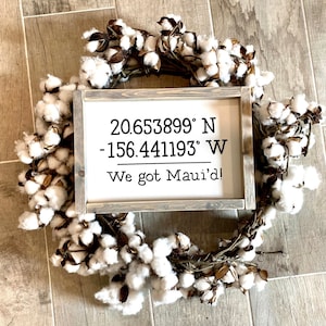 May include: A white wooden sign with the coordinates 20.653899&deg; N -156.441193&deg; W and the text "We got Maui'd!" The sign is framed in a rustic wood frame and is surrounded by a cotton wreath.