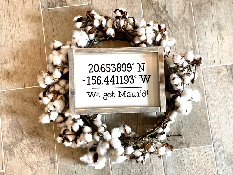 May include: A white wooden sign with the coordinates 20.653899&deg; N -156.441193&deg; W and the text "We got Maui'd!" The sign is framed in a rustic wood frame and is surrounded by a cotton wreath.