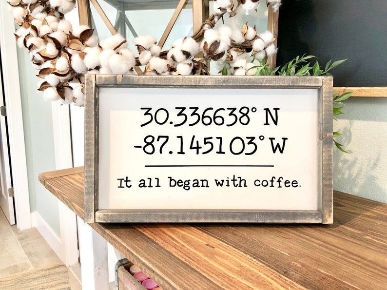 May include: A wooden sign with a distressed gray finish. The sign features white text that reads "30.336638&deg; N -87.145103&deg; W It all began with coffee."
