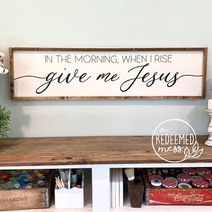 May include: Wooden sign with the text "IN THE MORNING, WHEN I RISE give me Jesus" in black cursive lettering. The sign is hanging on a light green wall above a wooden table.