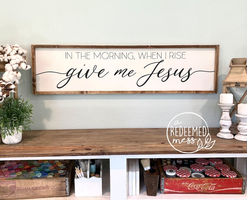 May include: Wooden sign with the text "IN THE MORNING, WHEN I RISE give me Jesus" in black cursive lettering. The sign is hanging on a light green wall above a wooden table.