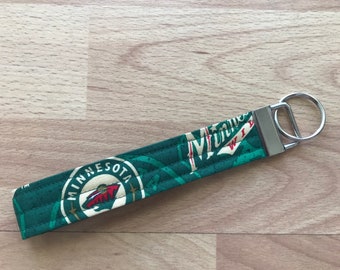 Pode incluir: Uma pulseira de chaveiro Minnesota Wild verde e branca. O tecido apresenta o logotipo e o nome da equipe. O chaveiro tem um fecho e um anel de metal prateado.