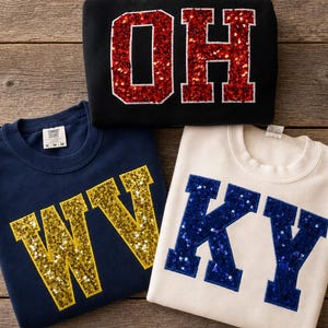 May include: Three crewneck sweatshirts featuring large varsity-style letters made of sequins. One black sweatshirt displays red 'OH' letters, one navy blue sweatshirt shows gold 'WV' letters, and one cream sweatshirt features blue 'KY' letters.