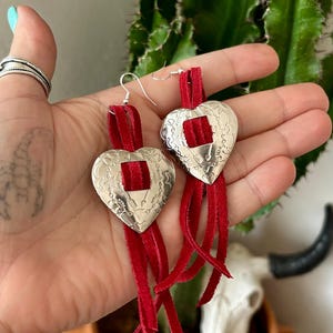 Könnte beinhalten: Silberne herzförmige Ohrringe mit quadratischem Ausschnitt und roter Wildlederfransen. Die Ohrringe haben ein Design im Western-Stil mit detaillierten Metallherzen. Rotes Wildleder sorgt für Farbe und Bewegung.