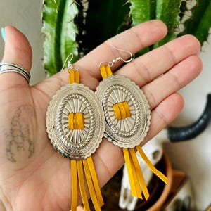 Könnte beinhalten: Silberfarbene ovale Ohrringe mit gewelltem Rand und einem quadratischen Mittelteil aus gelbem Leder. Die Ohrringe haben gelbe Lederfransen und werden an silbernen Haken getragen. Ein Accessoire im Western-Stil.