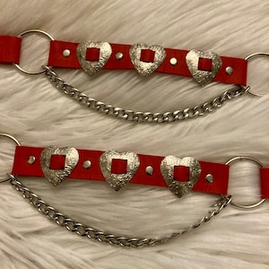 May include: Two red leather belts with silver heart-shaped buckles and silver chains attached. The belts have small silver studs along the edges.