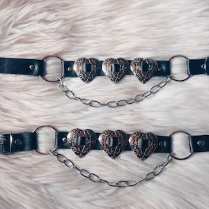Puede incluir: Dos cinturones de cuero negro con cadenas plateadas y detalles metálicos en forma de corazón. Cada cinturón tiene tres corazones.