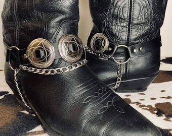 May include: A pair of black leather cowboy boots featuring intricate embossed patterns on the shafts and pointed toes. Each boot is adorned with a black leather harness strap, silver-toned circular conchos with star motifs, and a silver chain.
