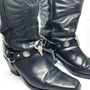 May include: A pair of black leather boots featuring a square toe, a tall shaft with stitched sunburst patterns, and a removable harness strap. The strap is adorned with silver-toned metal bull skull ornaments, circular studs, and a small chain link.