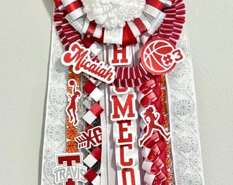 May include: A red and white homecoming mum featuring a basketball hoop, a basketball with #3, and the name Micaiah. It includes ribbons with basketball patterns, the word HOMECOMING, and the text Hoco 2026. The design incorporates glittery accents.
