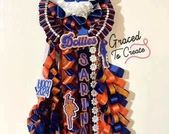 May include: A blue and orange graduation sash with a white flower, a white and blue "JM" emblem, and the words "Dollies", "Sarina", "Dance", and "2024".