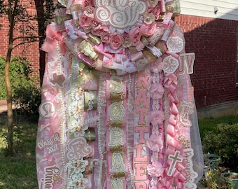 May include: A pink and gold graduation sash with a large floral wreath at the top. The sash has the name "Soraya" and the year "2023" on it. It also has a cross and the text "Proverbs 3:19".