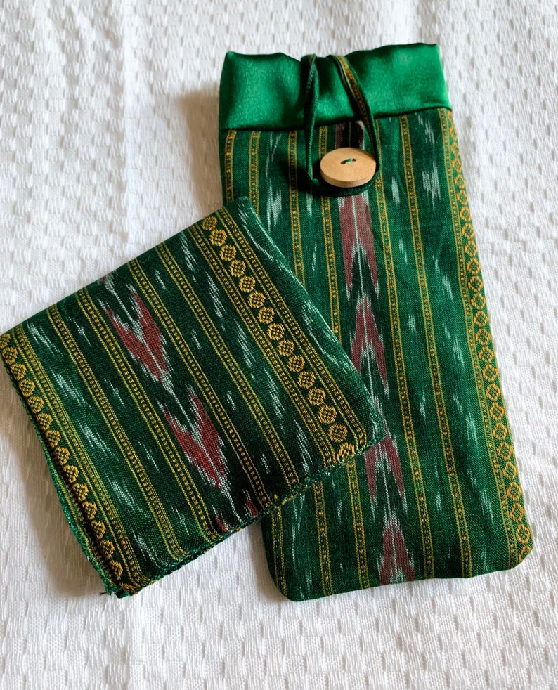 May include: A green and gold striped fabric pouch with a drawstring closure and a wooden button. The pouch is made of a soft, textured fabric and has a subtle ikat pattern.