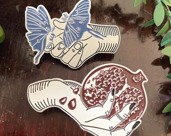 May include: Two enamel pins depicting hands holding a moth and a pomegranate. The moth is blue and white, and the pomegranate is red. Both pins are on a wooden surface.