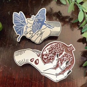May include: Two enamel pins depicting hands holding a moth and a pomegranate. The moth is blue and white, and the pomegranate is red. Both pins are on a wooden surface.